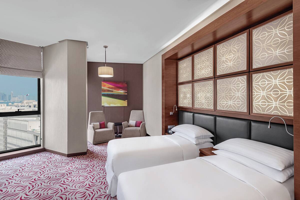 A hotel room with two beds, a city view, a decorative wall panel, and a modern chair.
