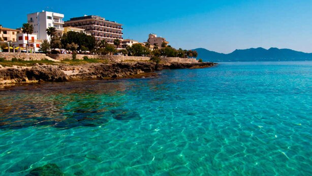 Hotel a la costa amb aigües blaves cristal·lines, un litoral rocos i edificis amb balcons davant del mar.