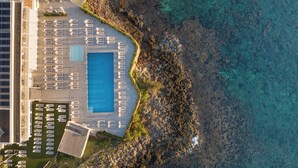 Vista aèria d'un complex turístic amb una gran piscina, gandules i una costa rocosa.