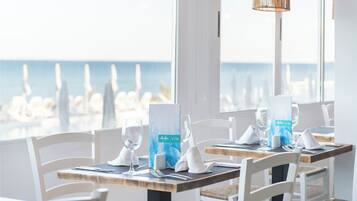 A dining area with tables set for two, white chairs, and a view of the ocean through large windows.