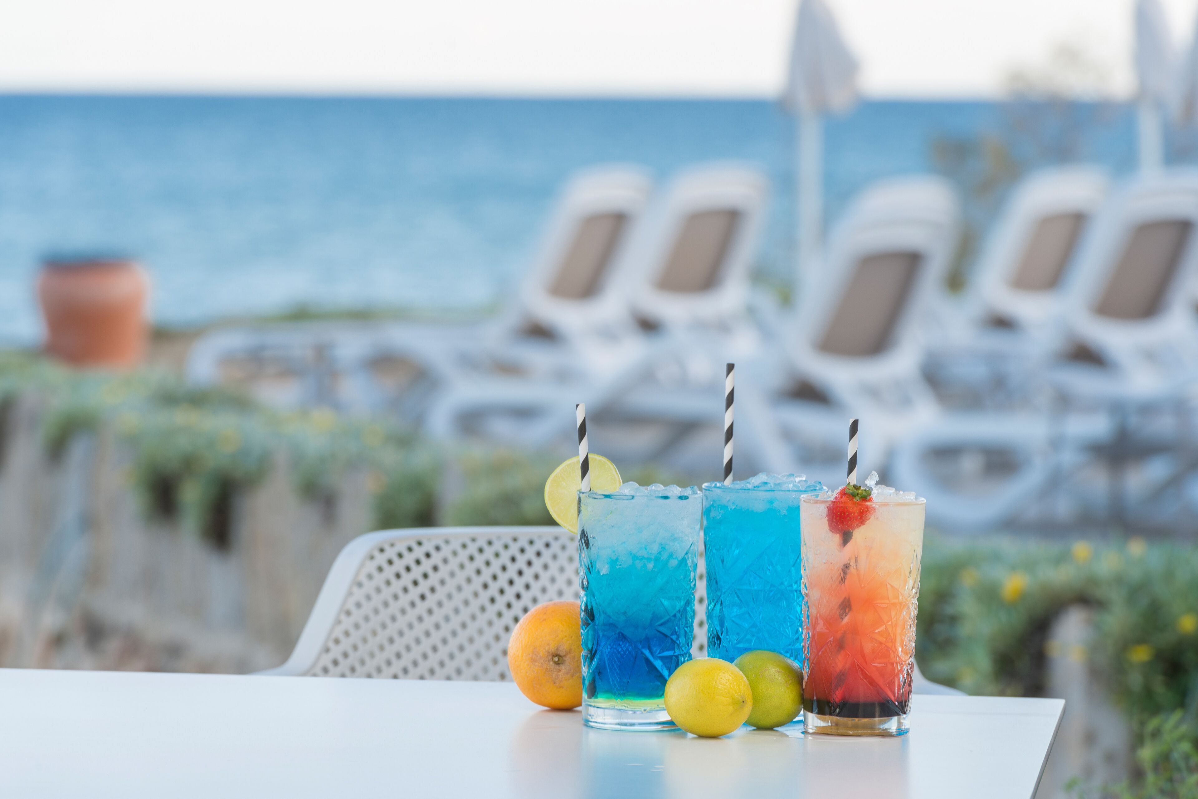 Three drinks on a table with a view of the sea and beach chairs in the background.