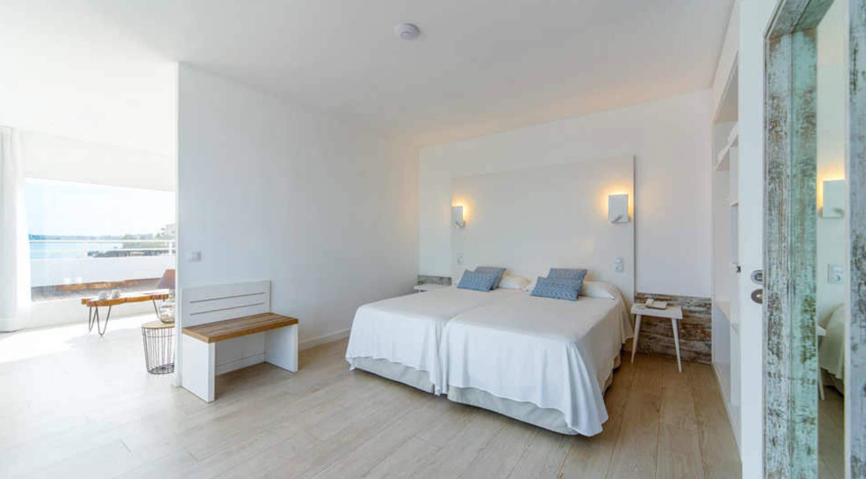 A modern bedroom with a large bed, a bedside table, a bench, and a view of the sea.