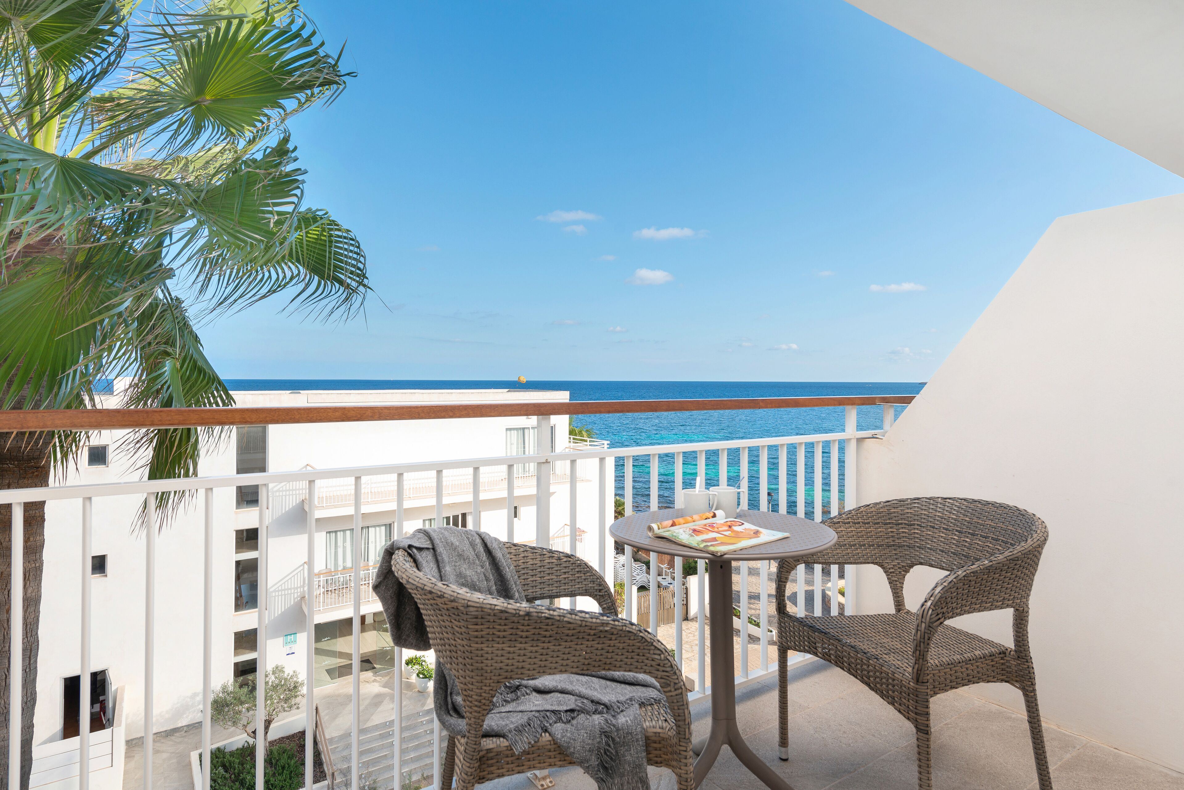 A balcony with wicker chairs and a table, overlooking a clear blue sea and a white building.