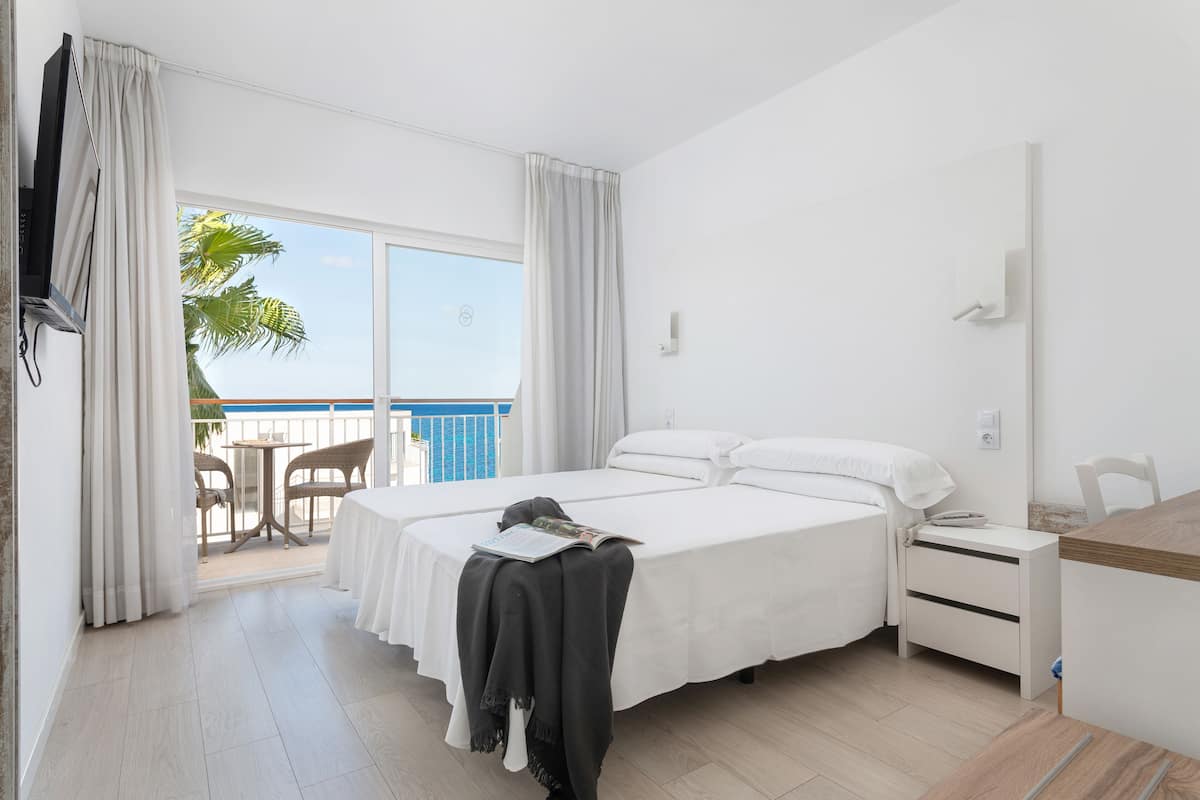 A hotel room with a bed, a bedside table, a chair, and a balcony with a view of palm trees.
