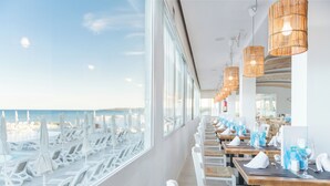 Un restaurant a la vora de la platja amb files de gandules i taules blanques, grans finestres amb vista a la platja i l'oceà, i llums penjants de vimbles.
