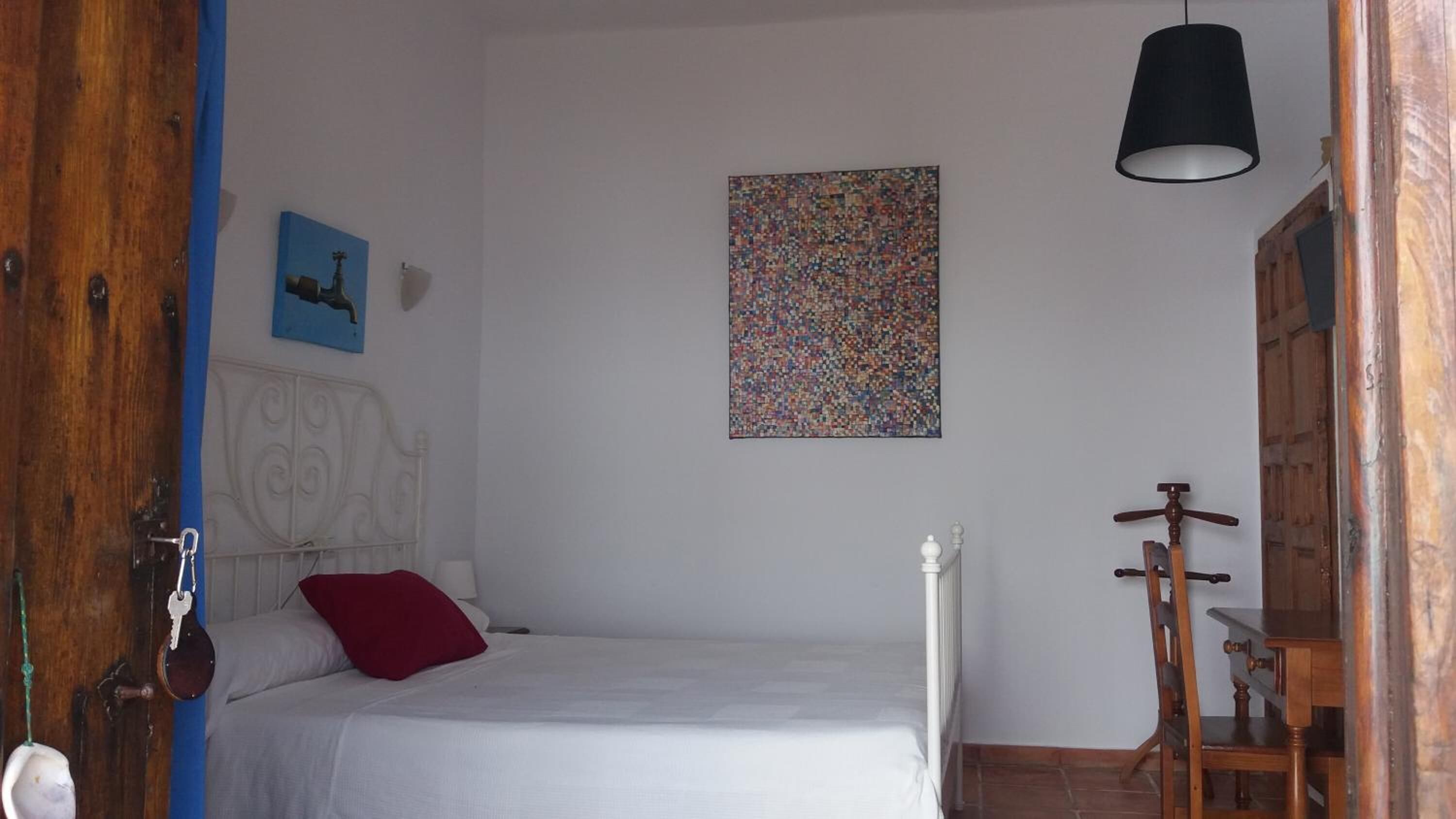 Un dormitorio con cama, almohada roja, cabecero blanco, puerta de madera, pared azul, radiador blanco y silla de madera, además de una lámpara negra.