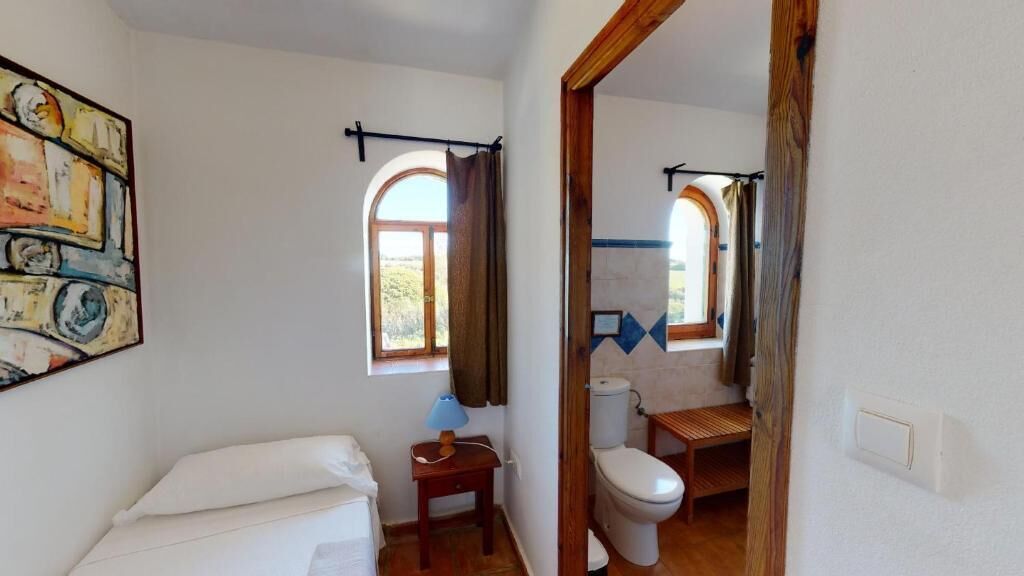 Ein Zimmer mit einem Bett, einem kleinen Tisch mit Lampe, einem Fenster mit Vorhängen und Blick auf ein Badezimmer mit Toilette und Waschbecken.