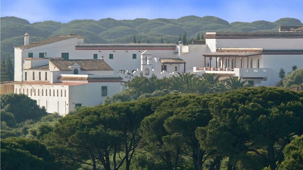 A large, white building with multiple sections, surrounded by dense greenery and hills in the background.