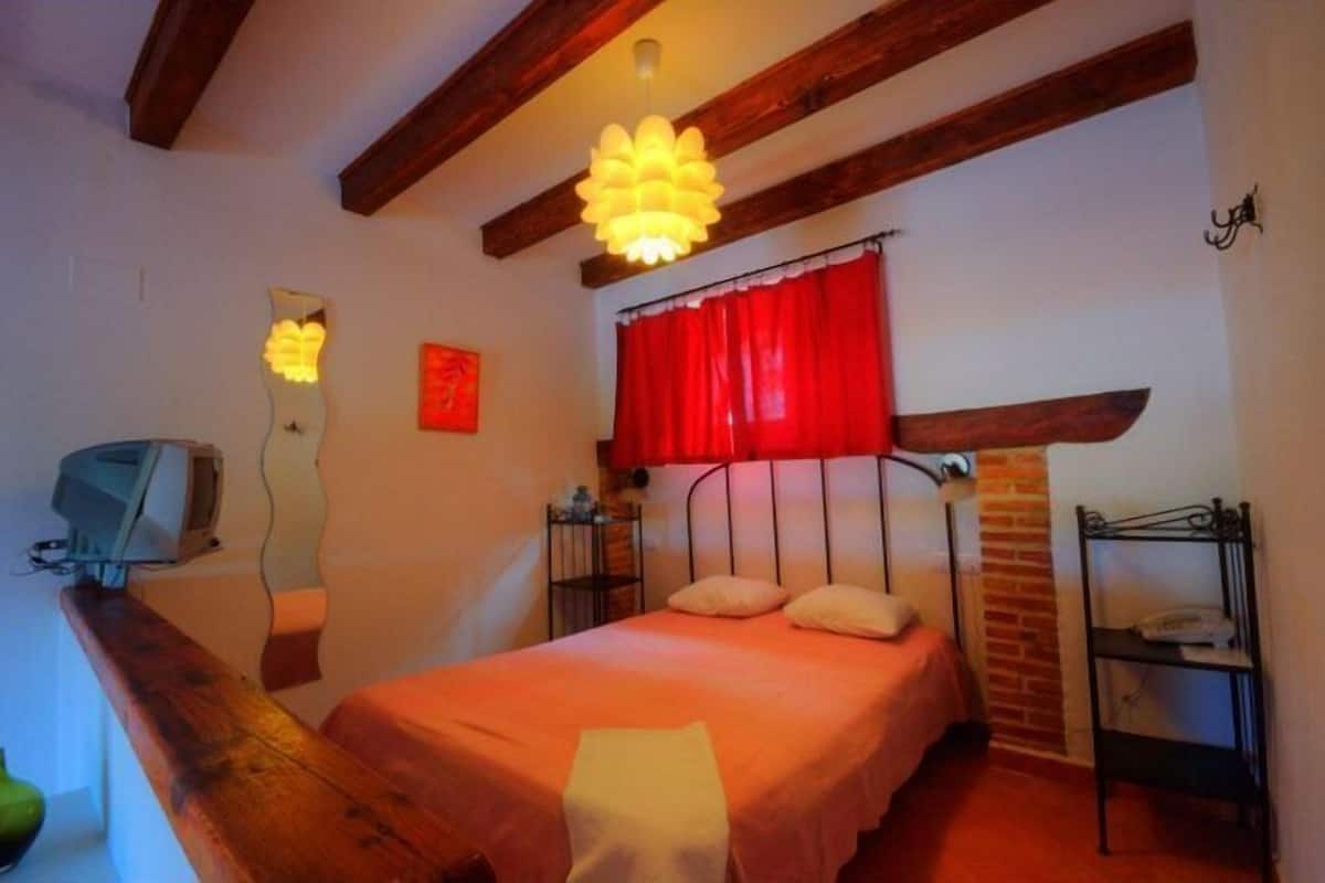 Un dormitorio con cama, vigas de madera, cortina roja, lámpara colgante, mesita con teléfono y televisor en la pared.