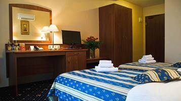 Habitación de hotel con escritorio, computadora, teléfono, espejo, cama con ropa de cama azul y blanca, y un florero con flores.