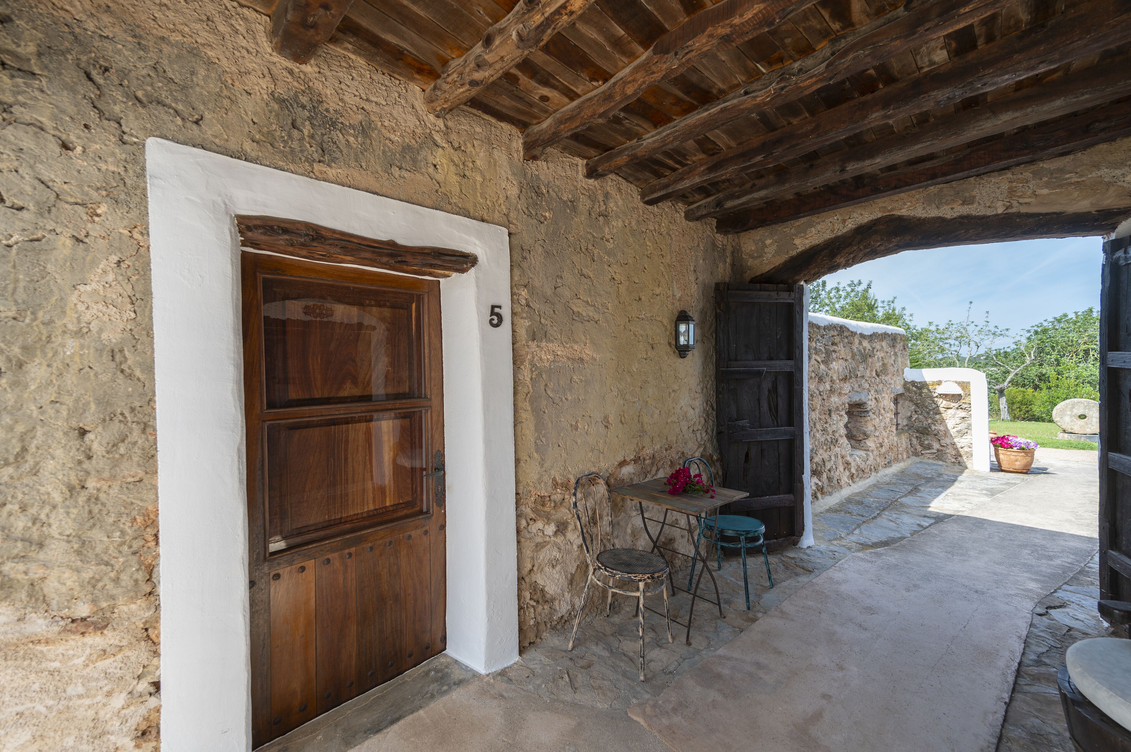 A rustic stone building with a wooden door marked with the number 5, a small table with a chair, and a potted plant.