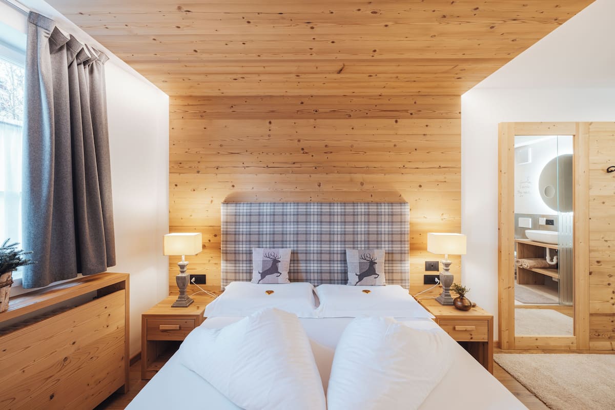 A bedroom with a wooden ceiling, a plaid headboard, two bedside tables with lamps, a large mirror, and a window with curtains.