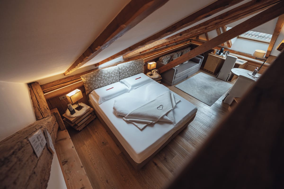 A bedroom with a wooden ceiling, a bed with white linens, a bedside table with a lamp, and a small kitchenette area.