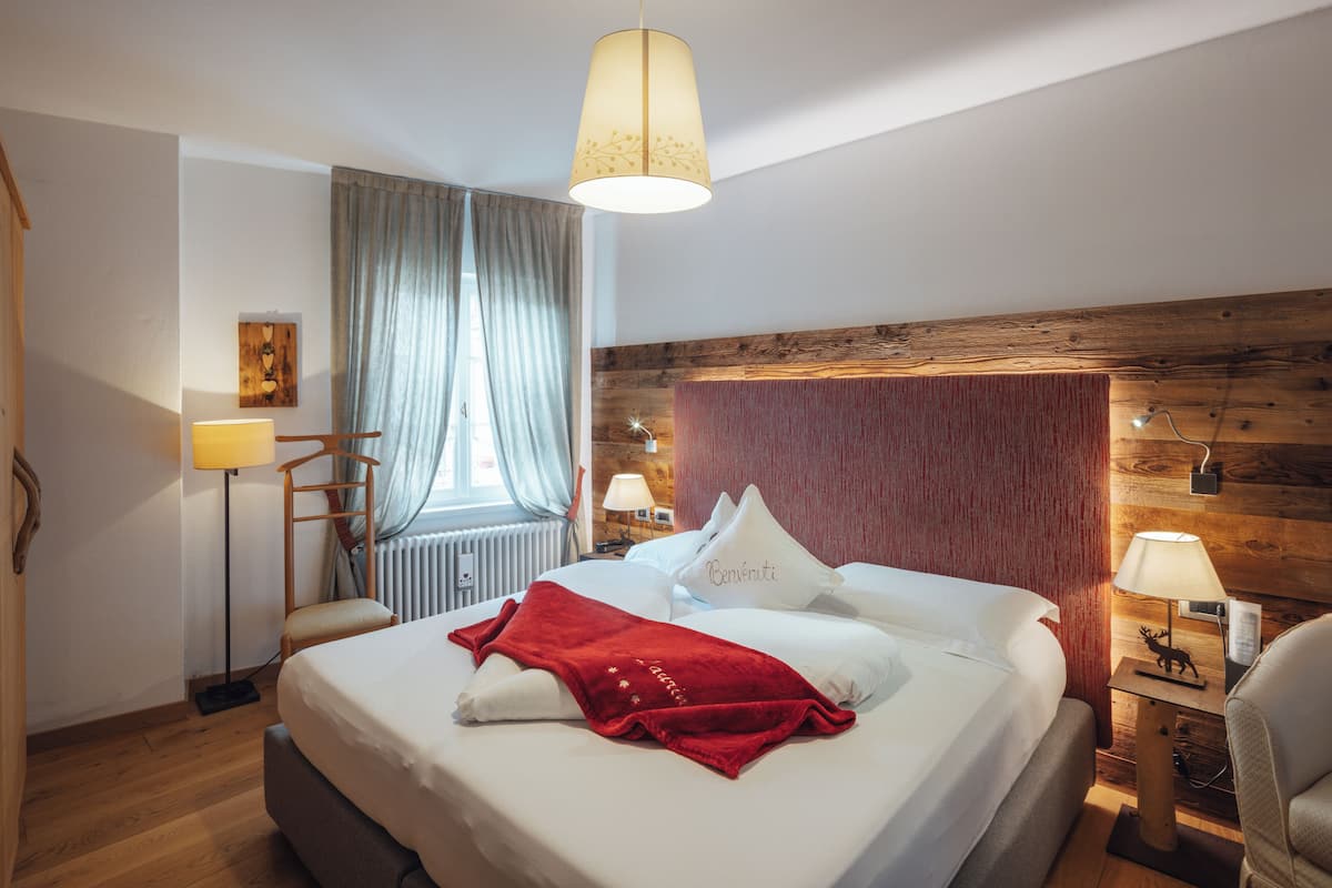 A hotel room with a large bed, a wooden headboard, a red throw blanket, and a window with curtains.