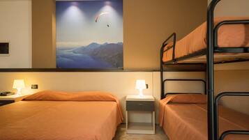 A hotel room with two beds, a bunk bed, a nightstand with a lamp, and a wall-mounted painting of a paragliding scene.