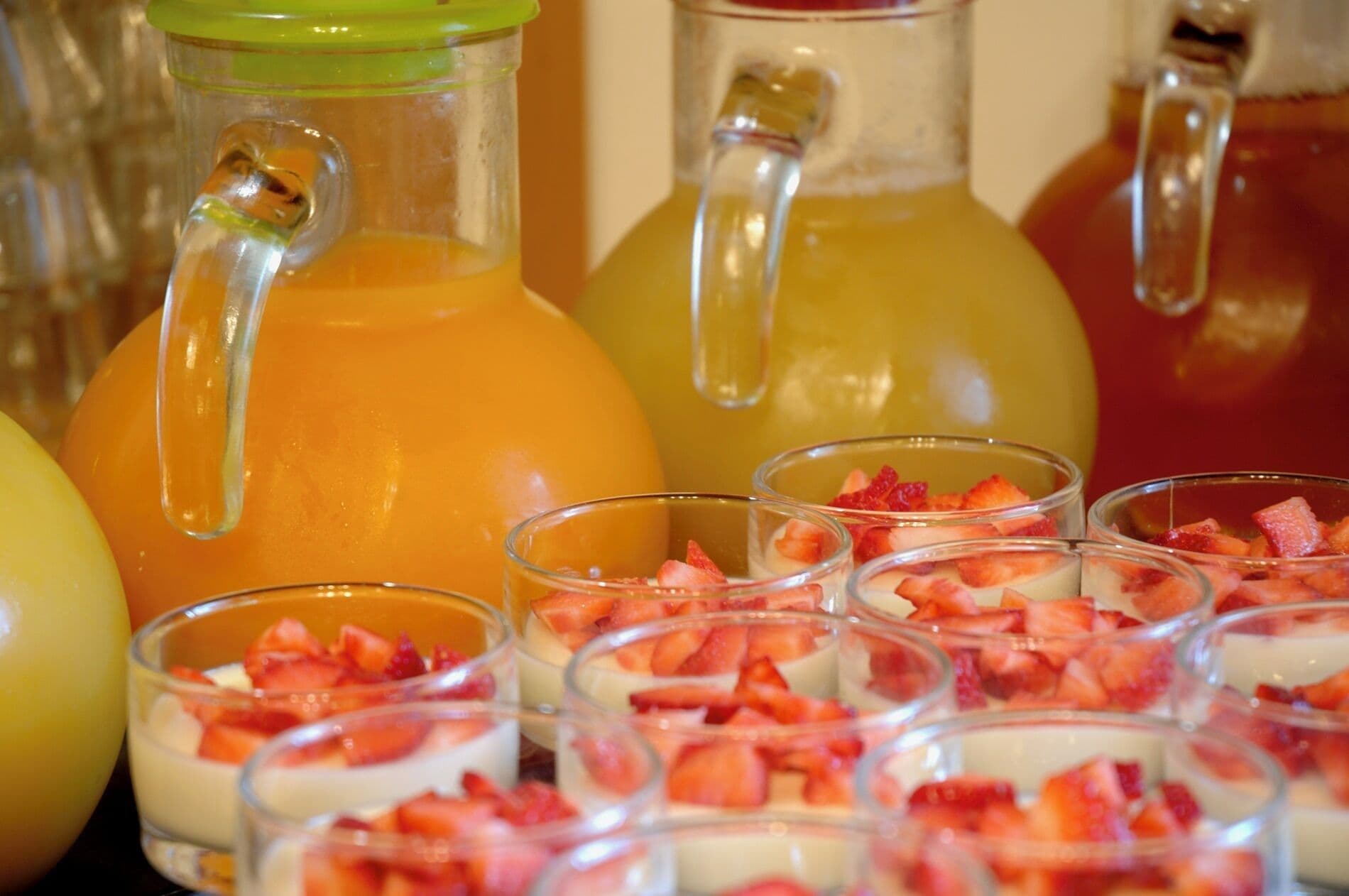 Una estación de bebidas con jarras y vasos, algunos con jugo de naranja y otros con una bebida amarilla, y pequeñas tazas adornadas con fresas.