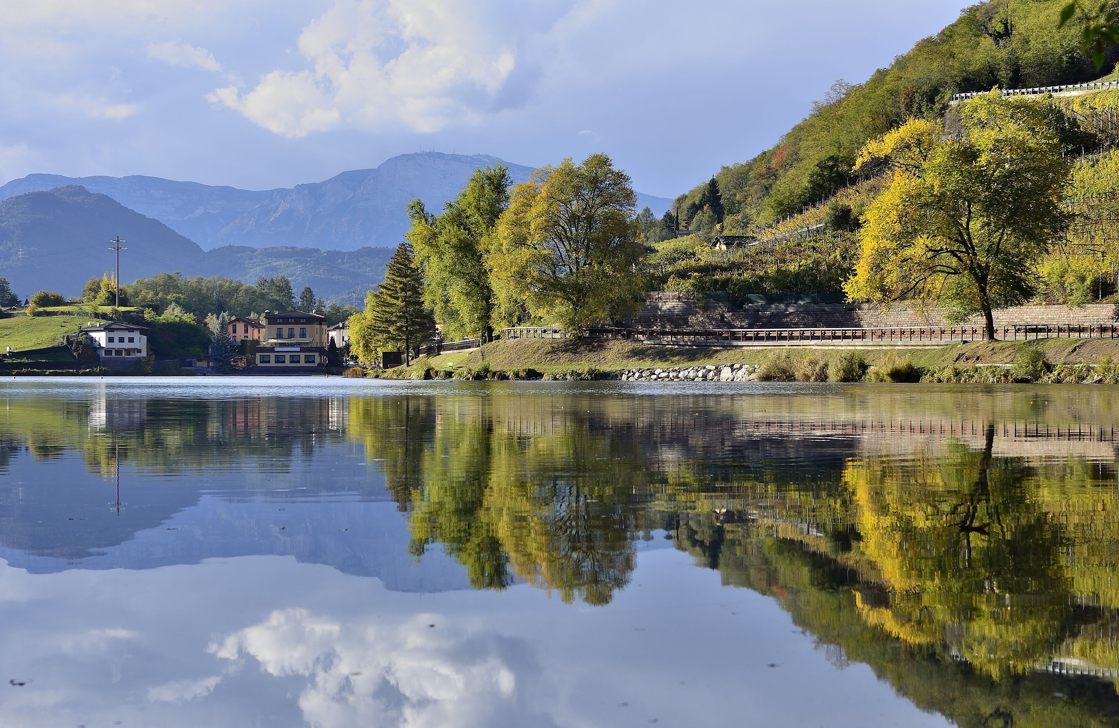 A serene lakeside view with a clear reflection of trees and buildings, a calm water surface, and a backdrop of mountains.