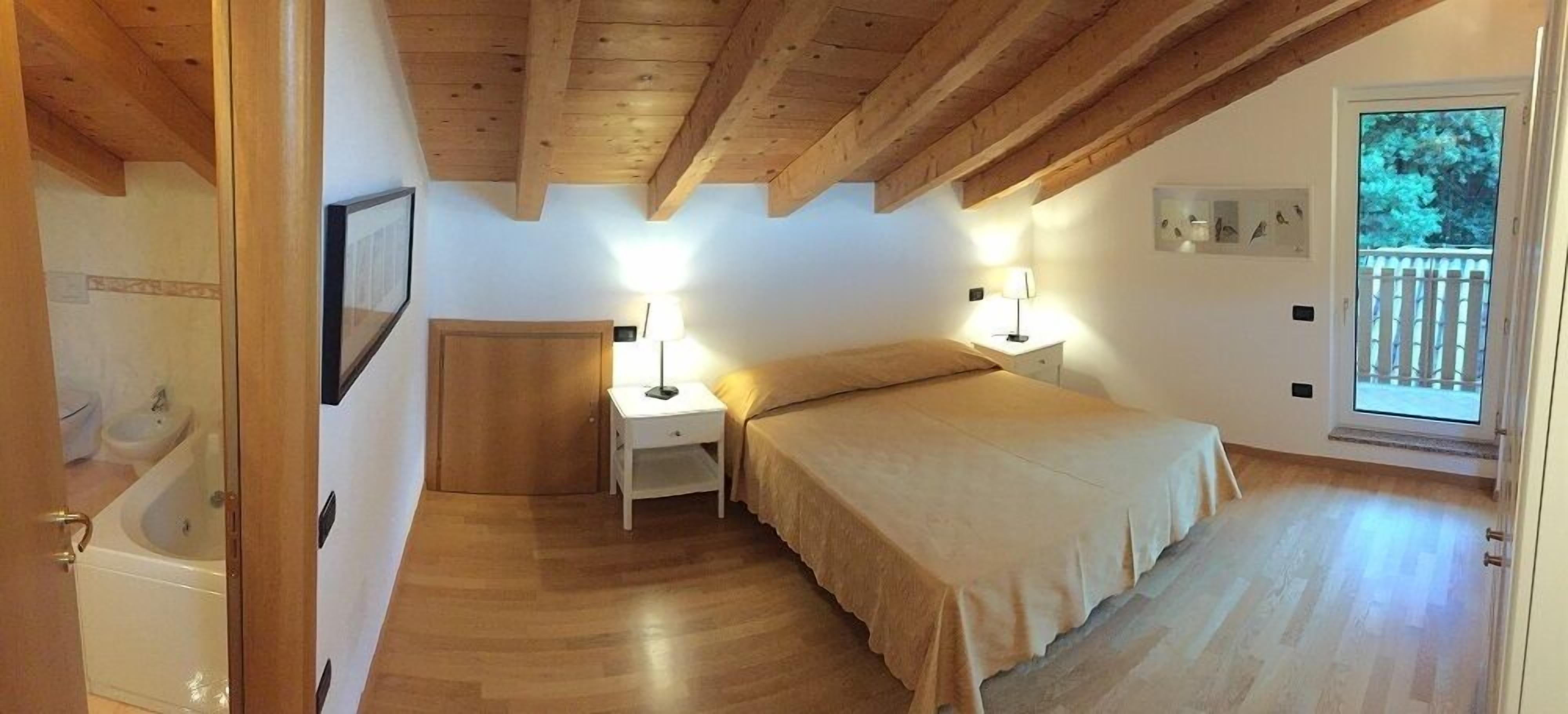 Un dormitorio con techo de madera, una cama con colcha color beige, dos mesitas de noche con lámparas, una ventana con vistas a árboles, y una puerta que da a un baño.