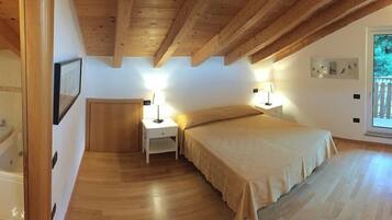 Un dormitorio con techo de madera, una cama con colcha color beige, dos mesitas de noche con lámparas, una ventana con vistas a árboles, y una puerta que da a un baño.
