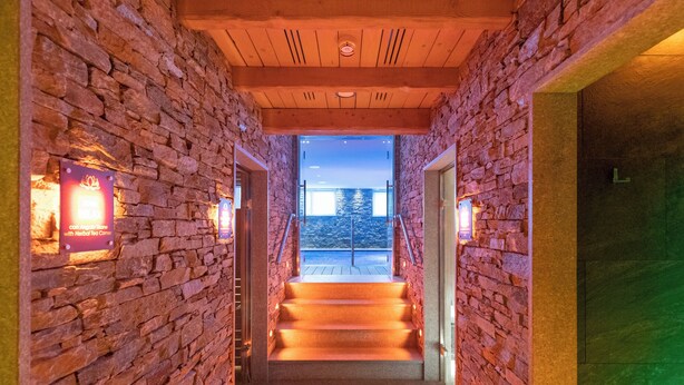 A stone-walled hallway with a wooden ceiling and a view of the outdoors at the end.