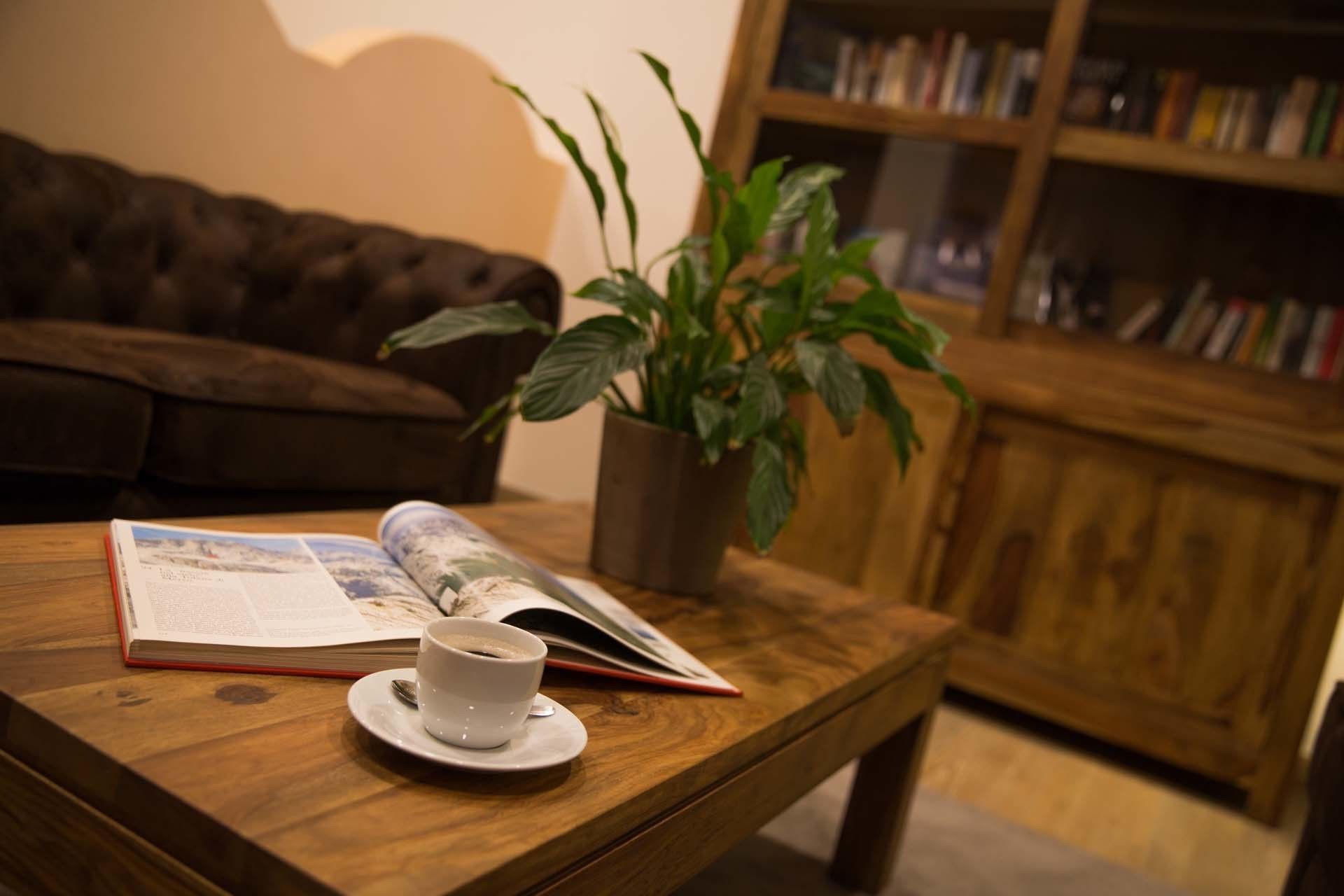 Una mesa de centro de madera con una taza blanca y una revista, una planta en maceta, un sofá marrón y una estantería de madera en una sala de estar acogedora.