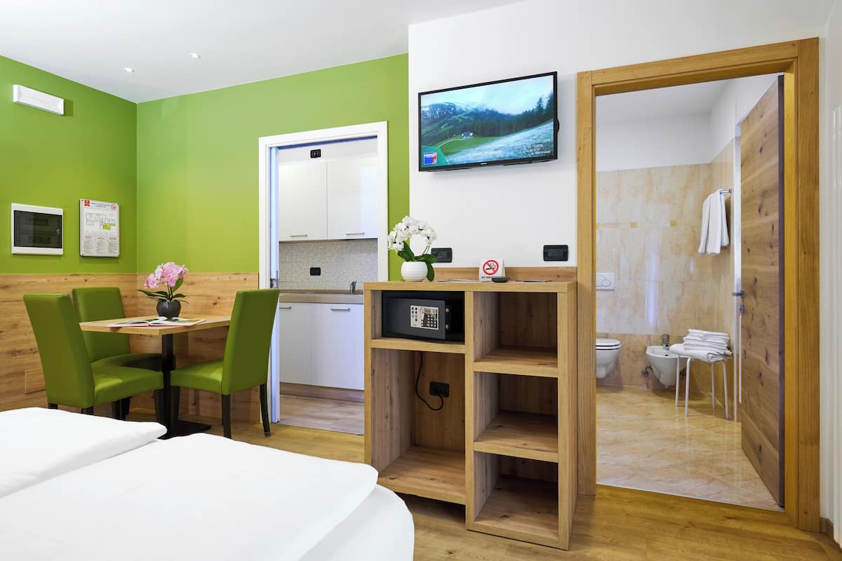 A hotel room with a bed, a desk with a computer, a chair, a small table with a vase, a TV on the wall, and a wooden door leading to a bathroom.