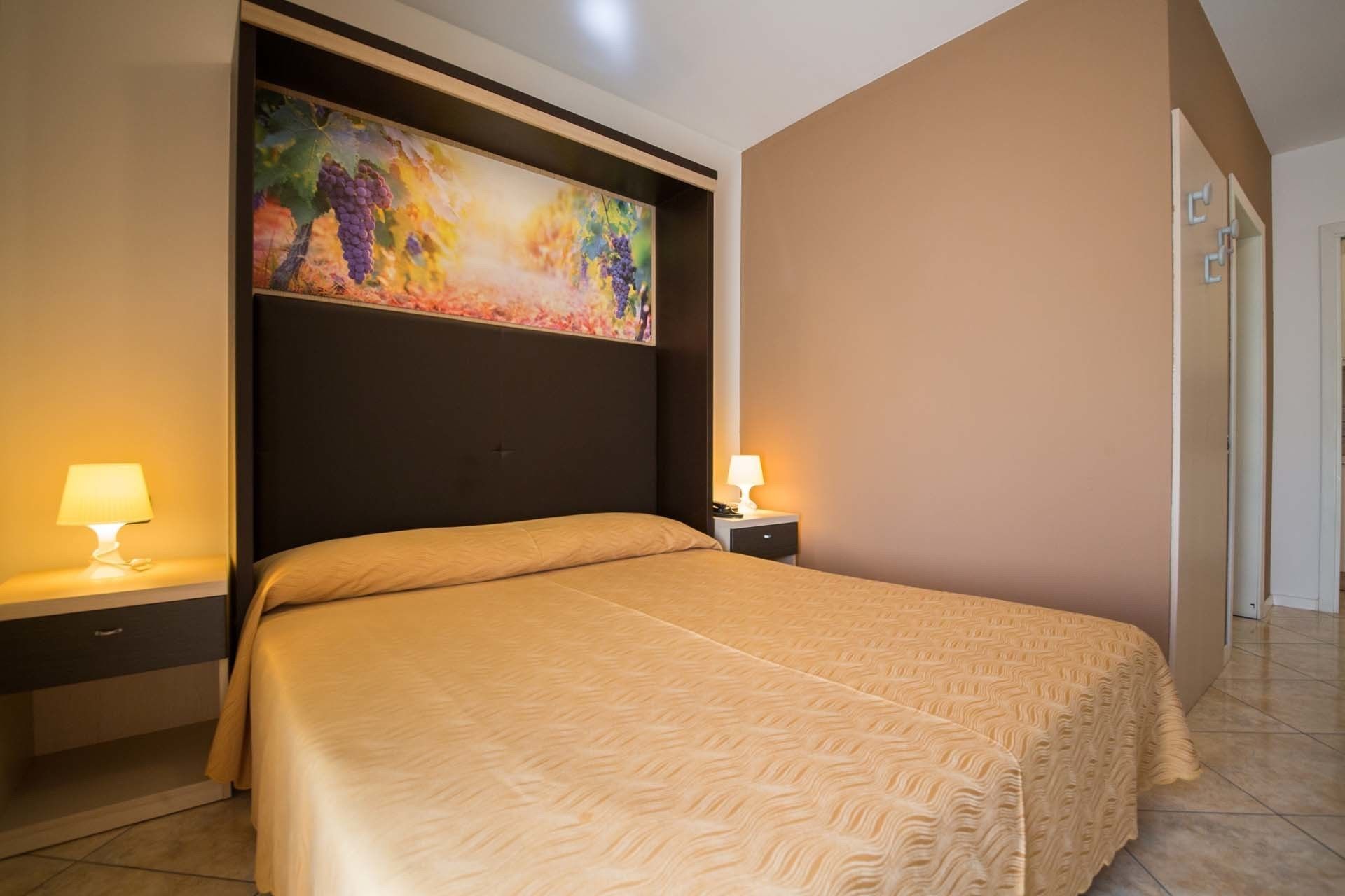 A hotel room with a large bed, two bedside tables with lamps, a headboard with a colorful painting, and a plain wall.