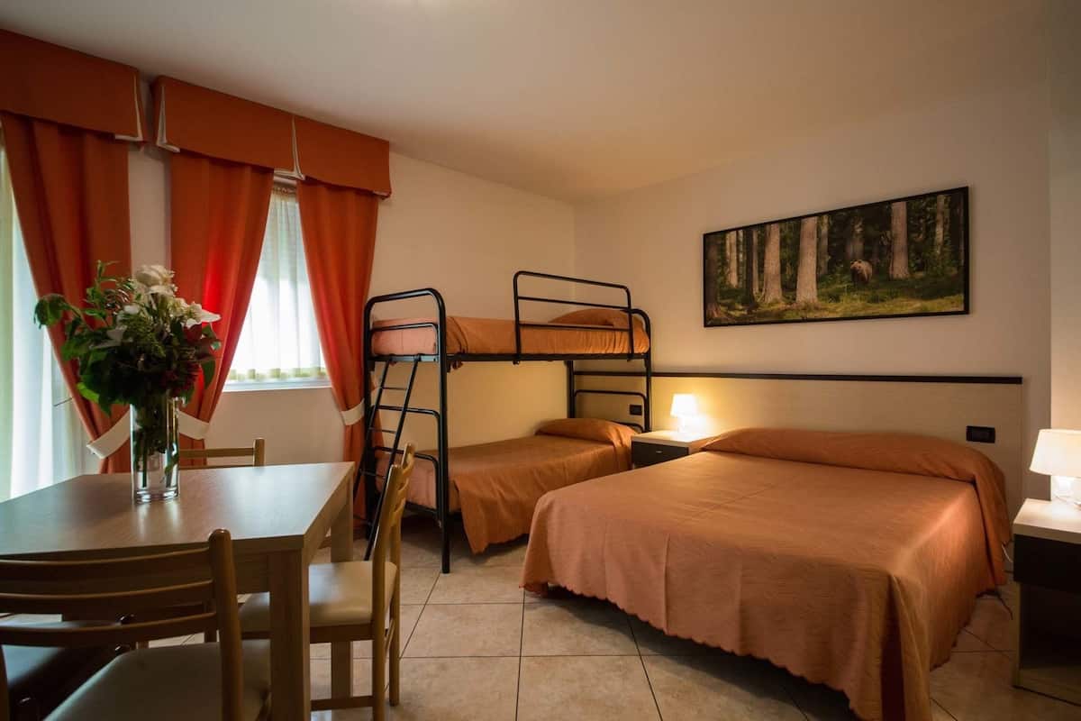 A dormitory room with bunk beds, a dining table with chairs, a vase with flowers, and a painting on the wall.