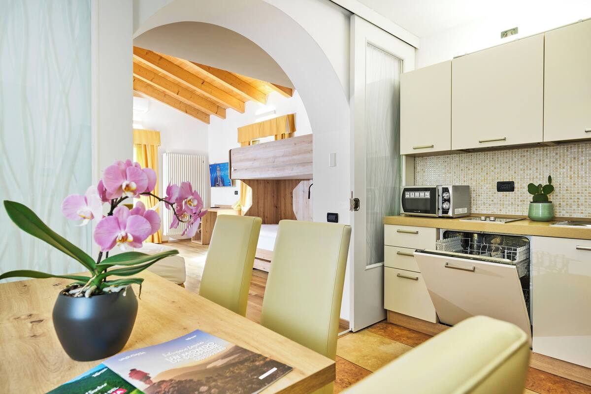 A modern kitchen with white cabinets, a microwave, and a dishwasher. There is a dining area with a wooden table and chairs, and a potted orchid on the table.