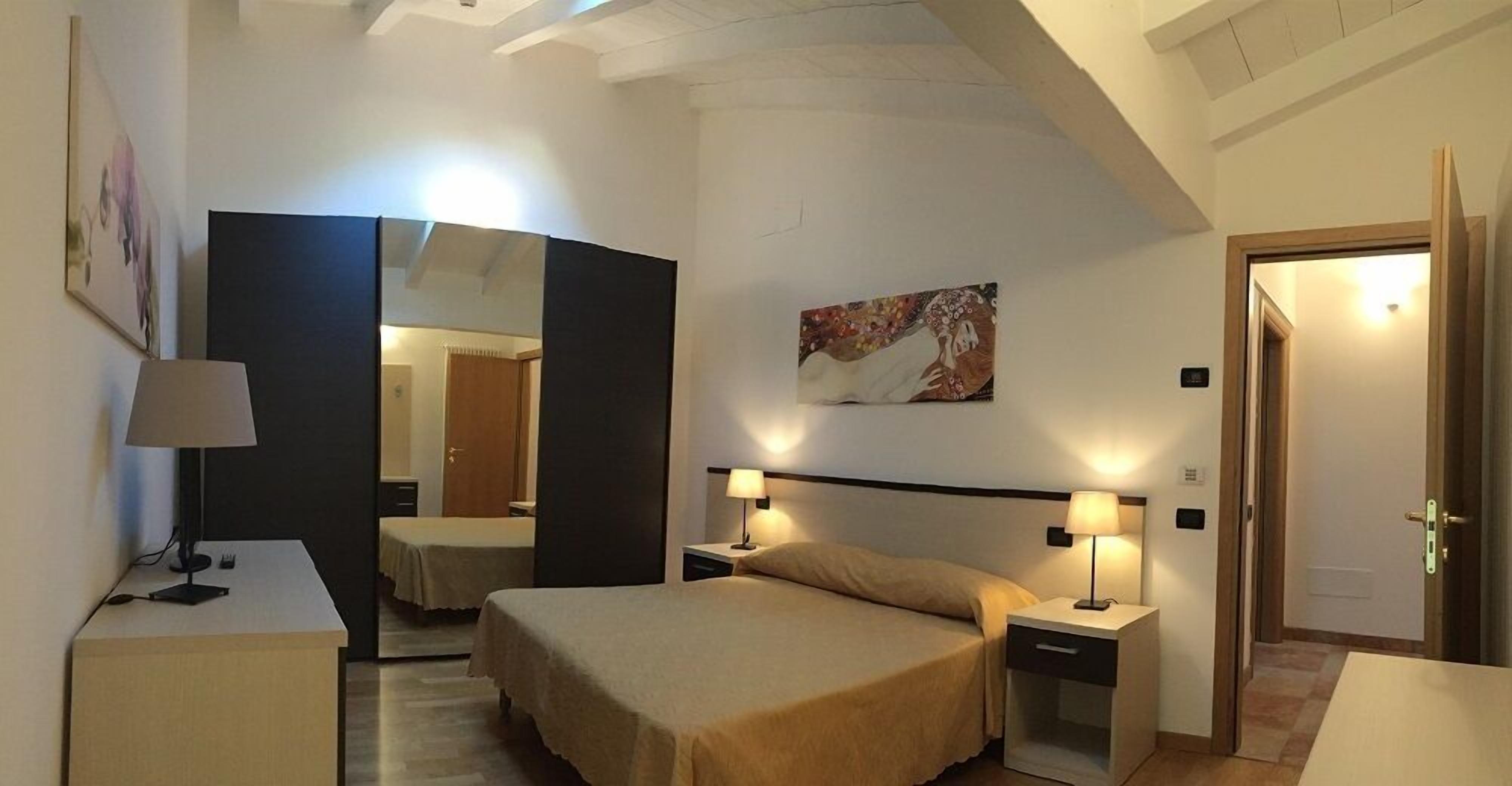 A hotel room with a bed, bedside tables, a desk, a lamp, a mirror, and artwork on the wall.