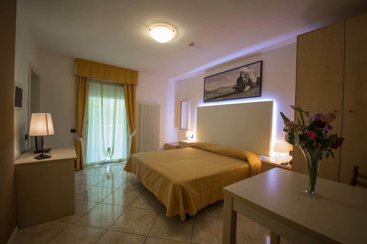 A hotel room with a bed, bedside tables, a desk, a chair, a radiator, a window with curtains, a painting on the wall, and a vase with flowers.