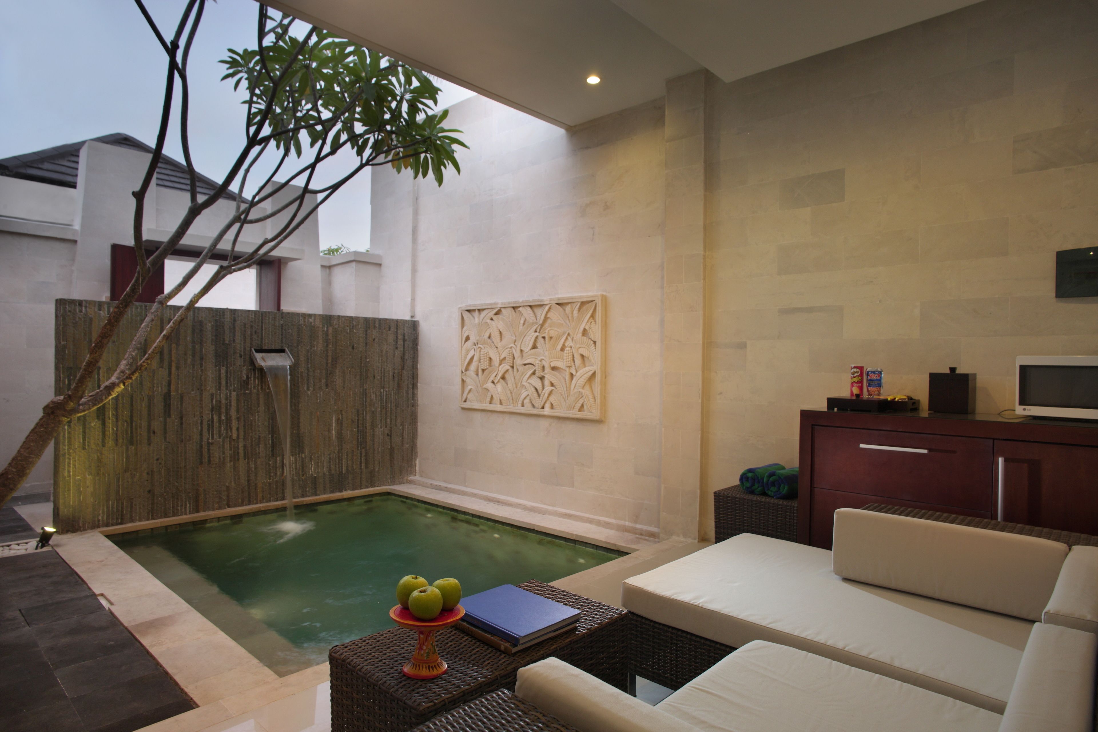A modern outdoor pool area with a lounge chair, a small table with a book and apples, and a decorative wall piece.