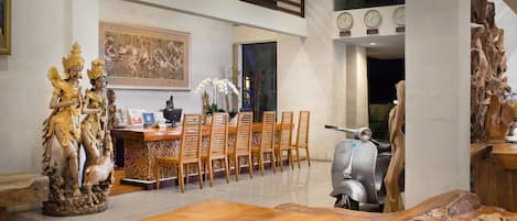 A dining area with a wooden table, chairs, and a statue. There is a scooter and a large wooden sculpture in the background.