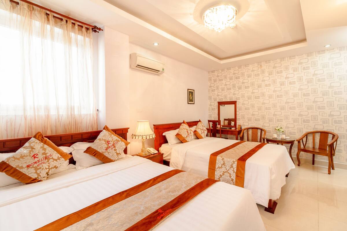 A hotel room with two beds, a desk, chairs, a lamp, and a ceiling light.