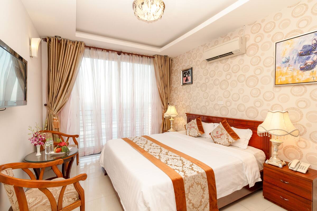A hotel room with a large bed, a bedside table with a lamp, a small table with flowers, and a TV on the wall.