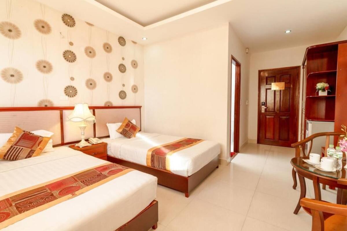 A hotel room with two beds, a wooden headboard, a bedside table with a lamp, a small table with a vase, and a wooden cabinet.