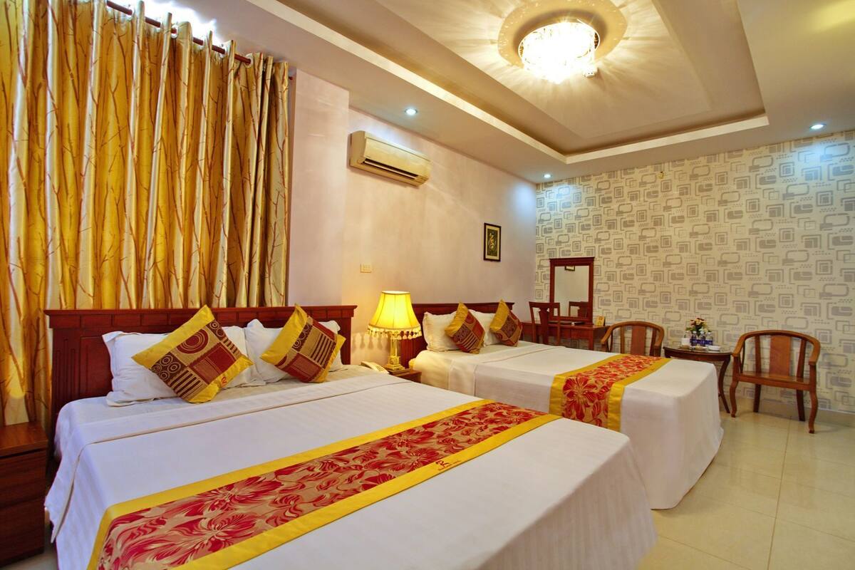 A hotel room with two beds, a desk, a chair, an air conditioning unit, and a patterned wallpaper.