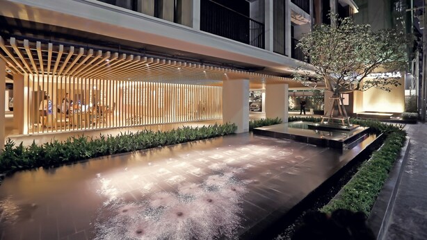 A modern building with a covered outdoor seating area, a water feature, and a tree.