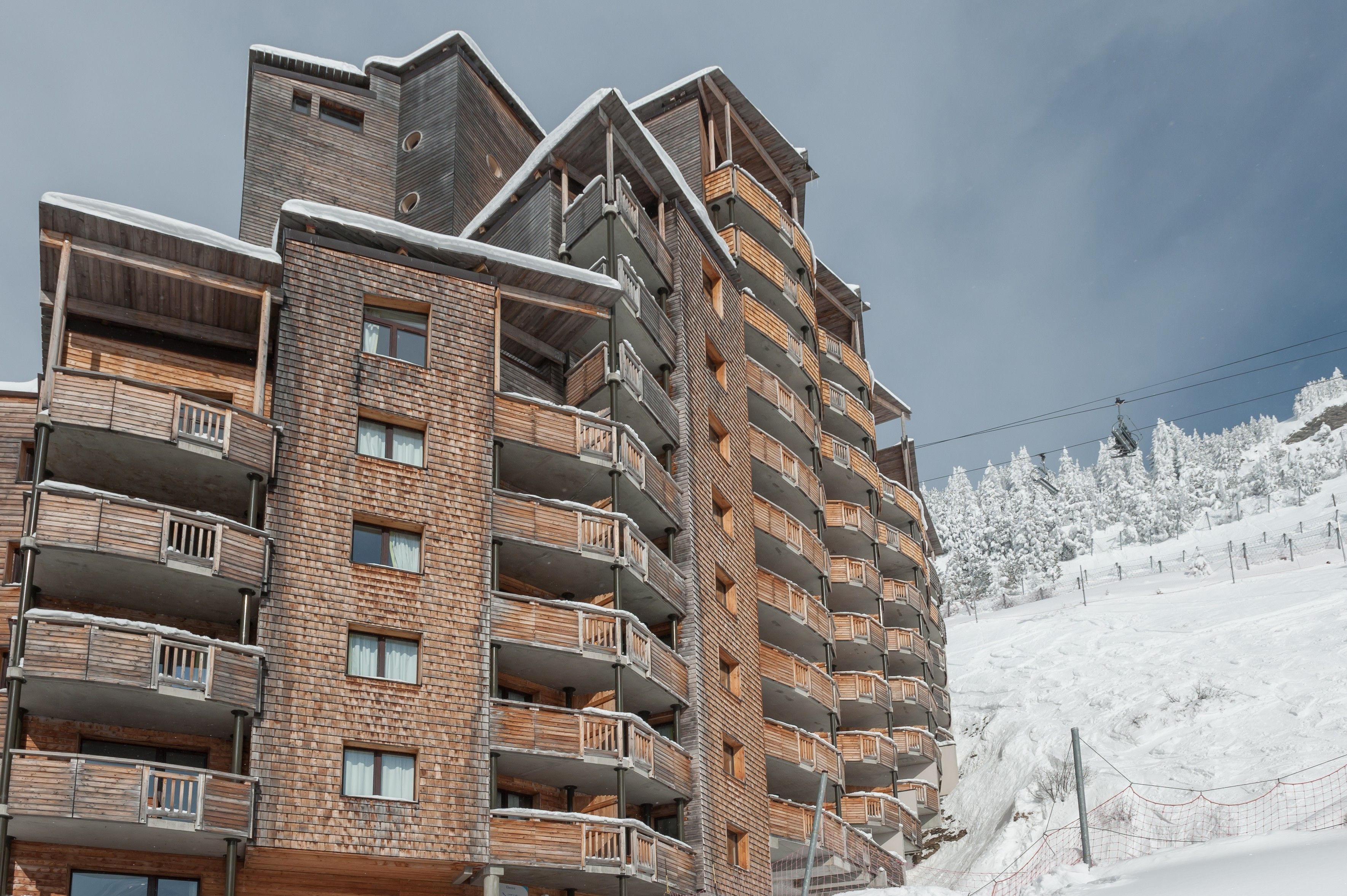 A multi-story building with balconies, likely a hotel or apartment complex, situated near a snowy slope.