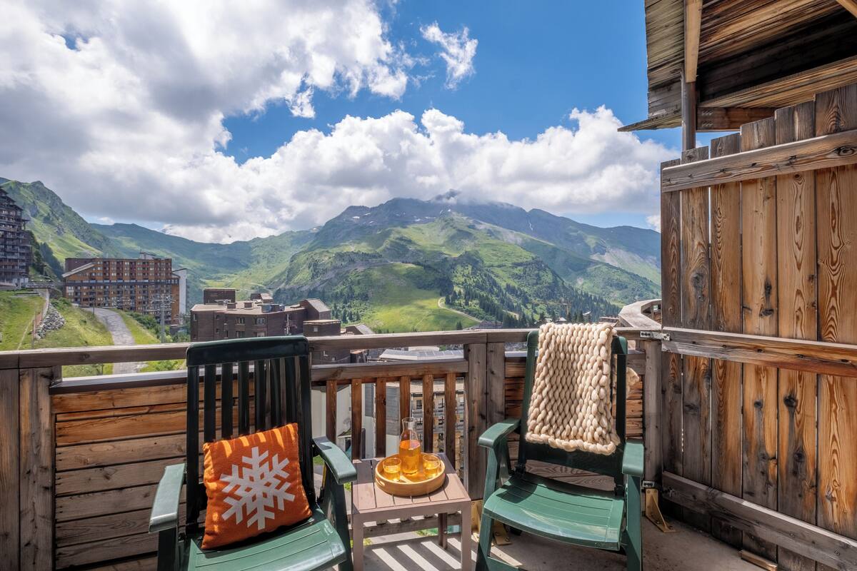 A wooden deck with two chairs, one with a snowflake pillow, and a tray with a drink. A mountain view is visible in the background.