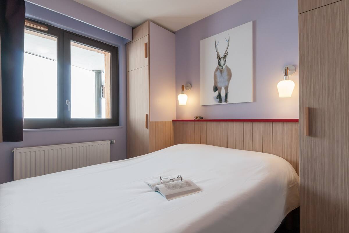 A hotel room with a large bed, a window with blinds, a wall-mounted light, and a framed picture of an animal.