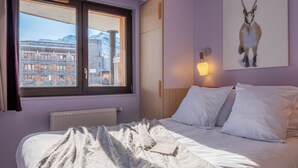 Un dormitorio con cama, ventana con vistas a edificios, una imagen de un animal colgada en la pared y una lámpara fijada a la pared.