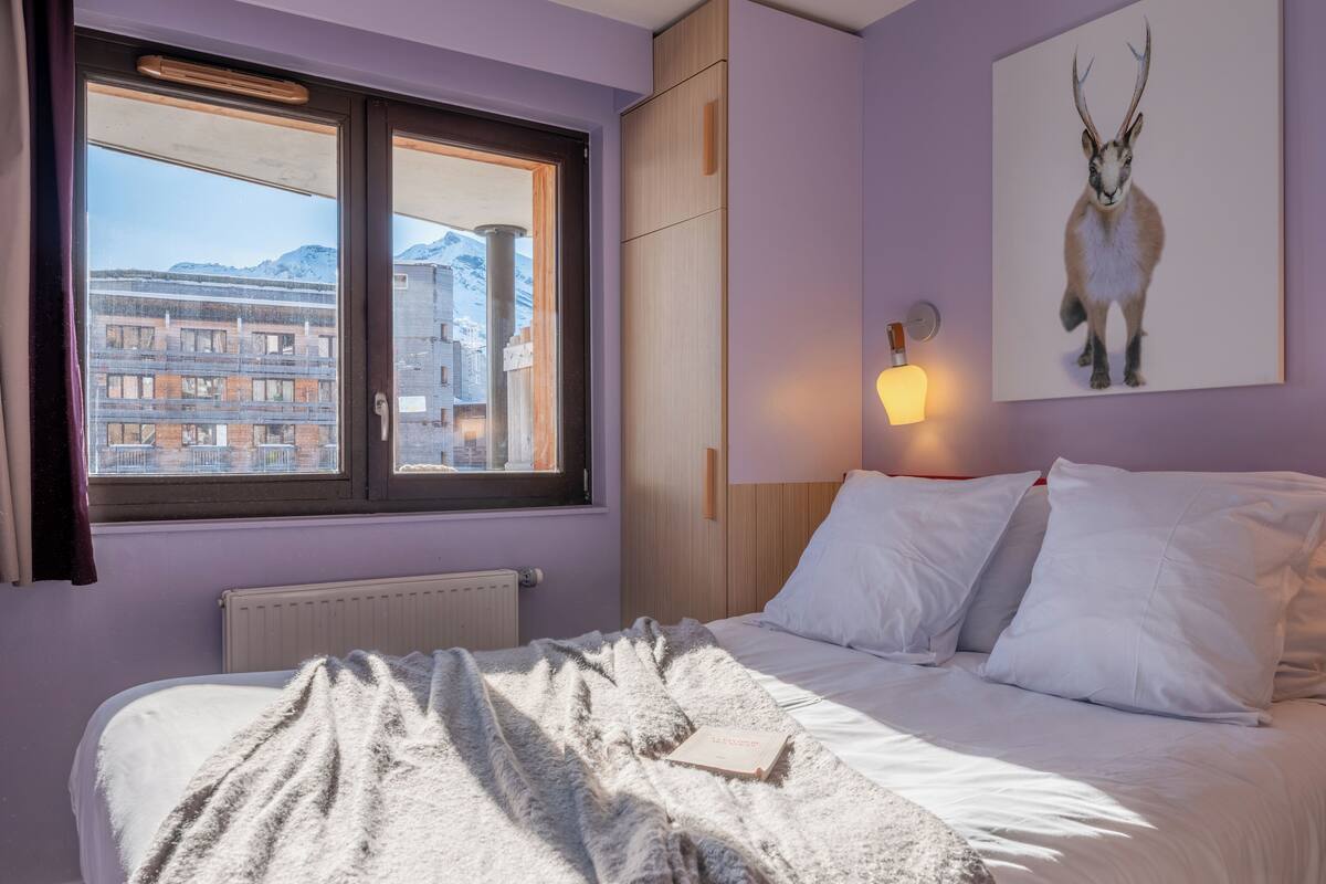 A bedroom with a bed, a window with a view of buildings, a mounted animal picture, and a wall-mounted light.