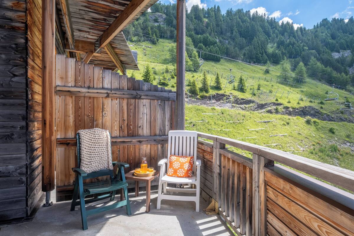 A wooden cabin with a balcony, a green chair, a white chair with an orange cushion, and a table with a drink and a tray.