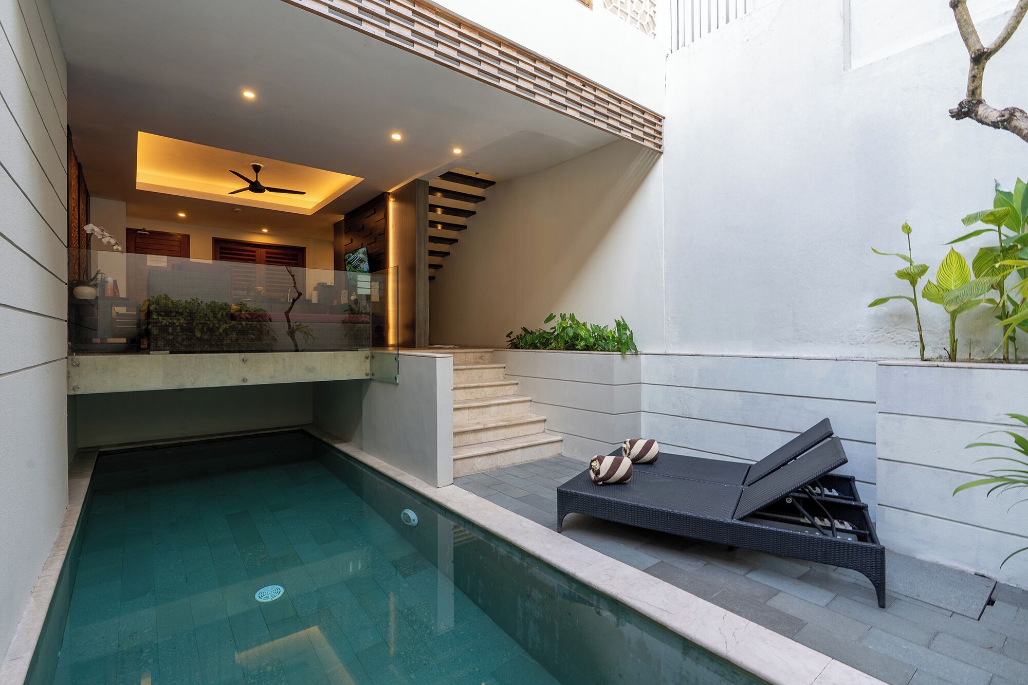 Un área de piscina moderna al aire libre con una silla de reposo, una escalera y un espacio con plantas cubierto de cristal.