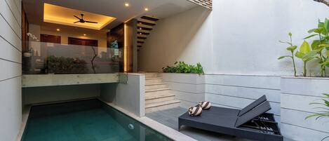 A modern outdoor pool area with a lounge chair, a staircase, and a glass-enclosed space with plants.