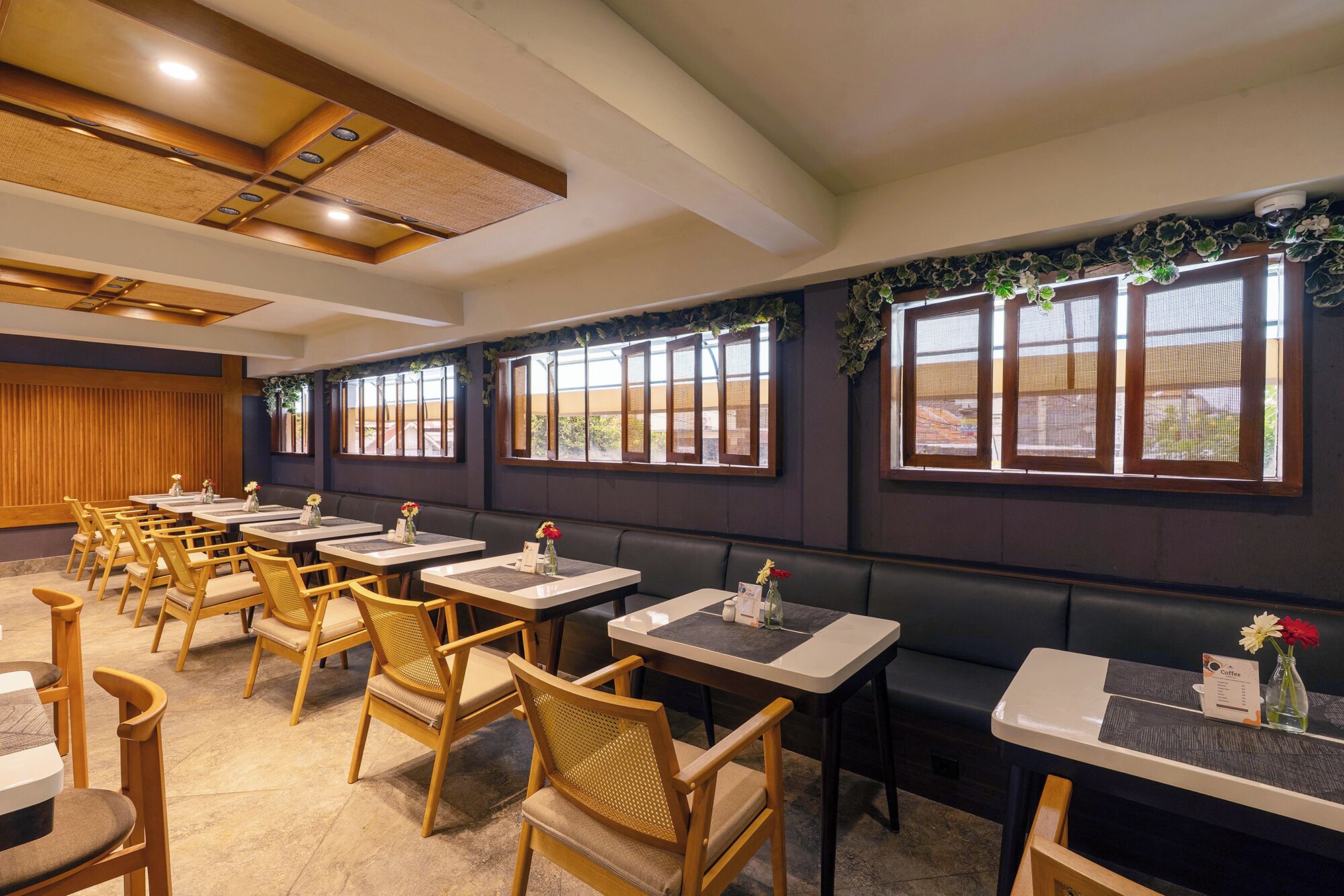 Een restaurant met houten stoelen en tafels, grote ramen met gordijnen en een plafond met inbouwverlichting.