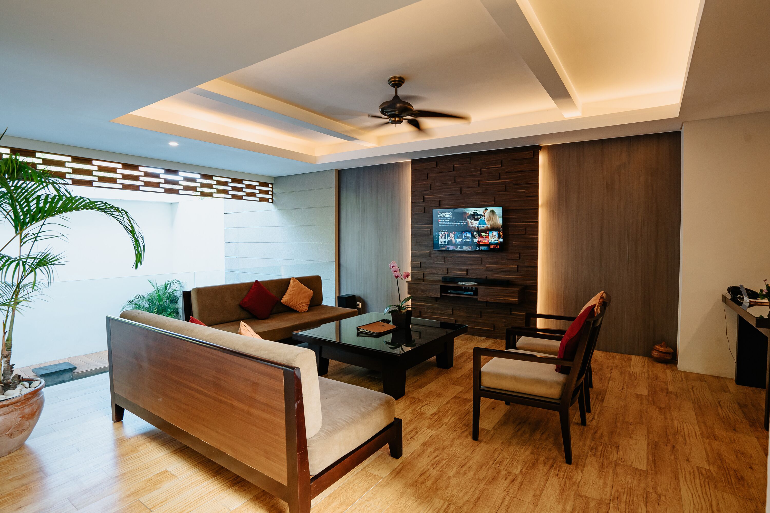 A modern living room with a sofa, coffee table, and chairs, a television mounted on a wooden wall, and a ceiling fan.