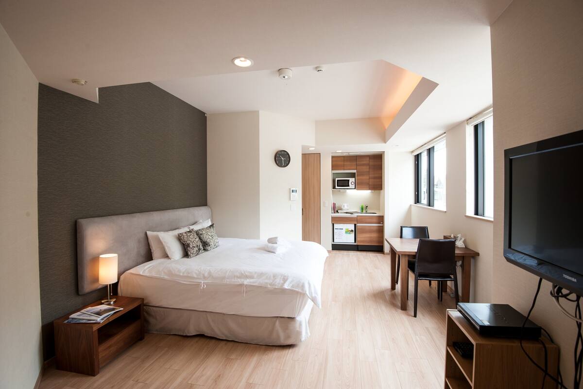 A modern hotel room with a large bed, a desk, a TV, and a kitchenette in the background.