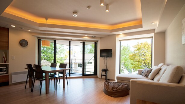 Uma sala de estar moderna com área de jantar, um sofá, uma TV de tela plana e uma janela grande com vista para árvores.